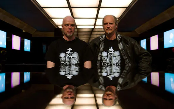 Uwe Büttner und Ralph Metz in einem dunklen Raum mit Bildschirmen, gespiegelt in einer glänzenden Tischplatte
